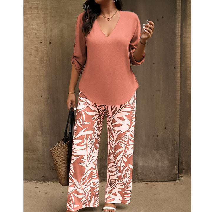 Woman in rust-colored V-neck top and rust-and-white leaf-print pants with a straw handbag against a rustic wall.