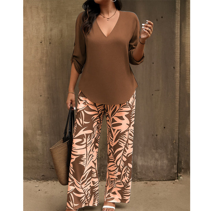 Woman wearing brown V-neck top and brown and peach leaf-print pants, carrying a straw handbag, standing by a textured concrete wall.