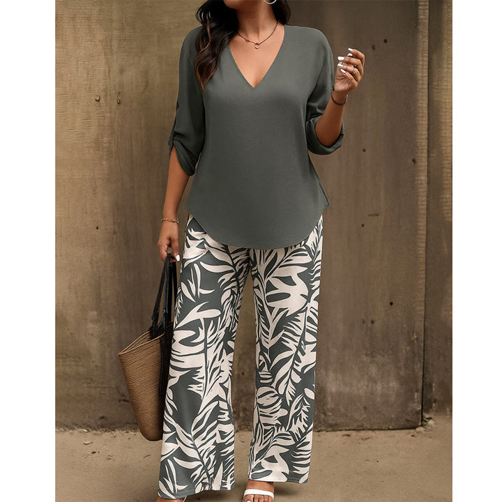 Woman dressed in gray V-neck top with gray and white leaf-print pants, holding a straw handbag, posed against a concrete background.