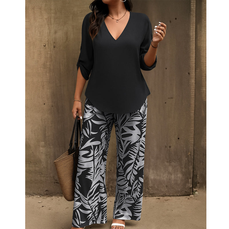 Woman in black V-neck top and black and white leaf-print pants holding a straw handbag against a textured wall.