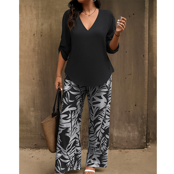 Woman in black V-neck top and black and white leaf-print pants holding a straw handbag against a textured wall.