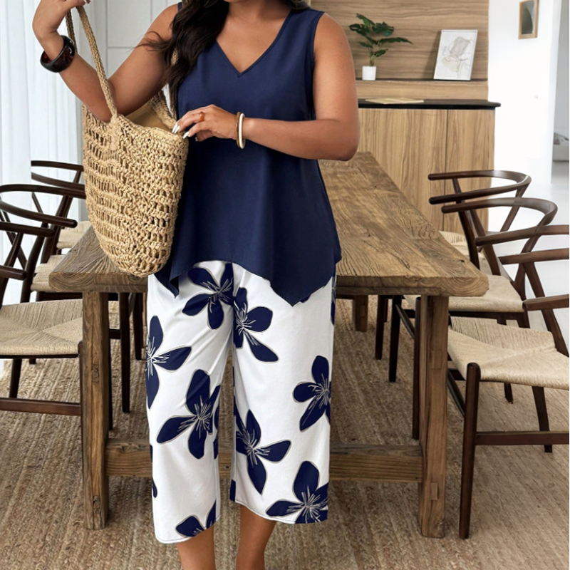 Woman in navy tunic and white floral pants holding a woven bag in front of a wooden dining table and chairs.