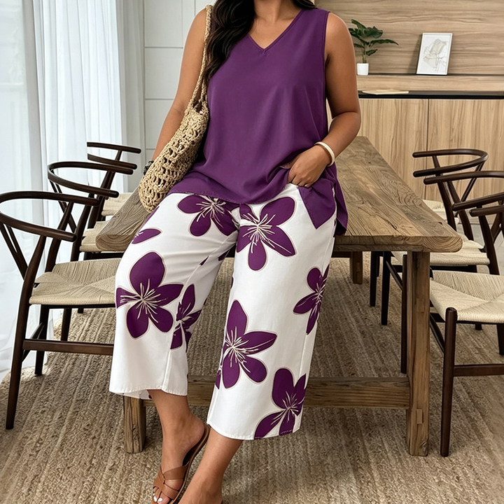 Woman sitting wearing a violet tunic and white floral pants holding a woven bag in a wooden furnished dining area.