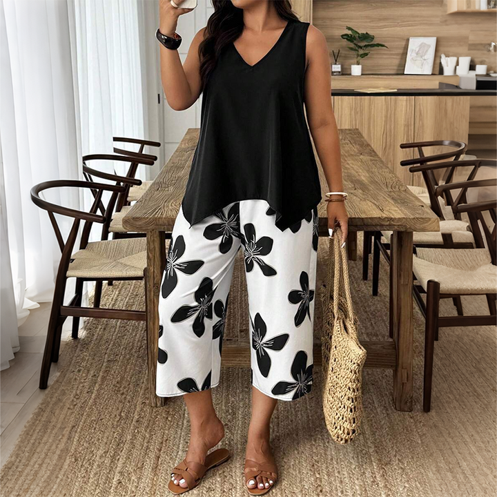 Woman in a black tunic and white floral pants with a woven bag standing in a modern wood-furnished dining room.