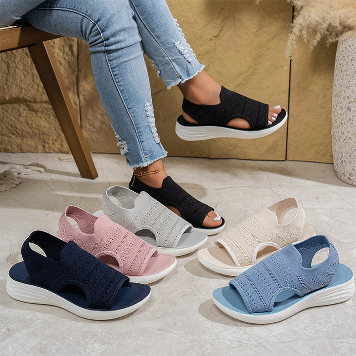 Assorted colors of knit platform sandals displayed on a textured floor, person wearing black pair seated on a chair.