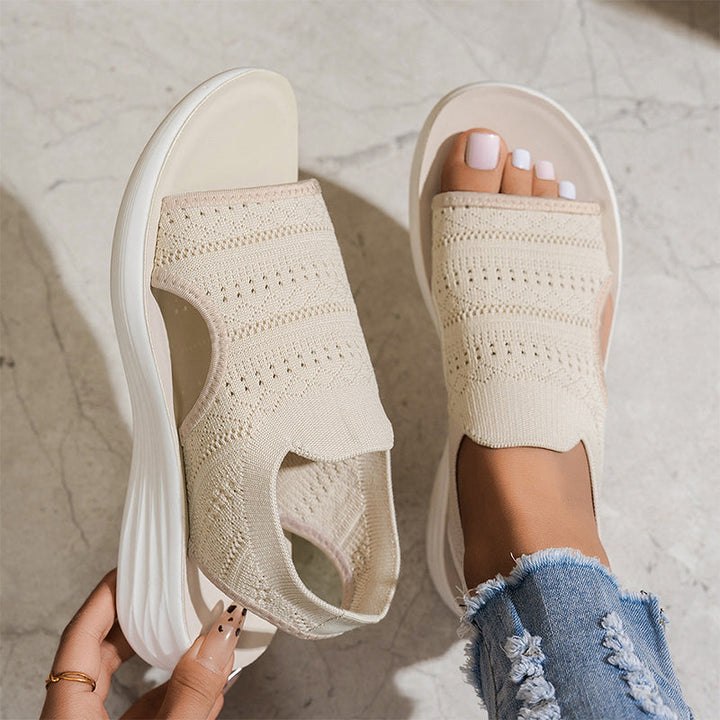 Beige breathable knit platform sandals with white soles on feet, one hand adjusting, on textured floor with distressed jeans.