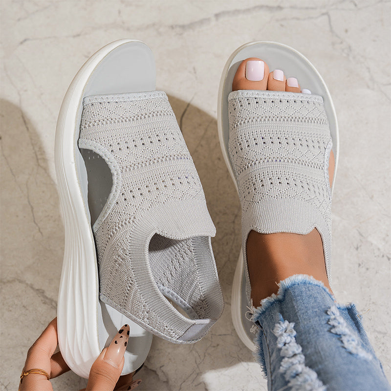 Gray breathable knit platform sandals with white soles on feet, one hand adjusting, on textured floor with distressed jeans.