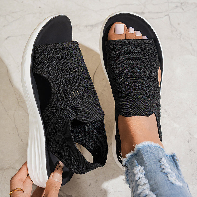 Black breathable knit platform sandals with white soles on feet, one hand adjusting, on textured floor with distressed jeans.
