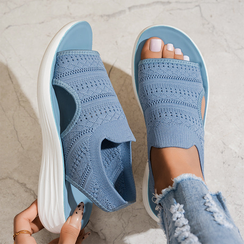 Blue breathable knit platform sandals with white soles on feet, one hand adjusting, on textured floor with distressed jeans.