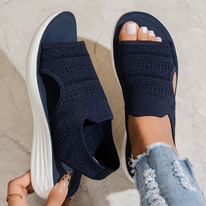Navy breathable knit platform sandals with white soles on feet, one hand adjusting, on textured floor with distressed jeans.