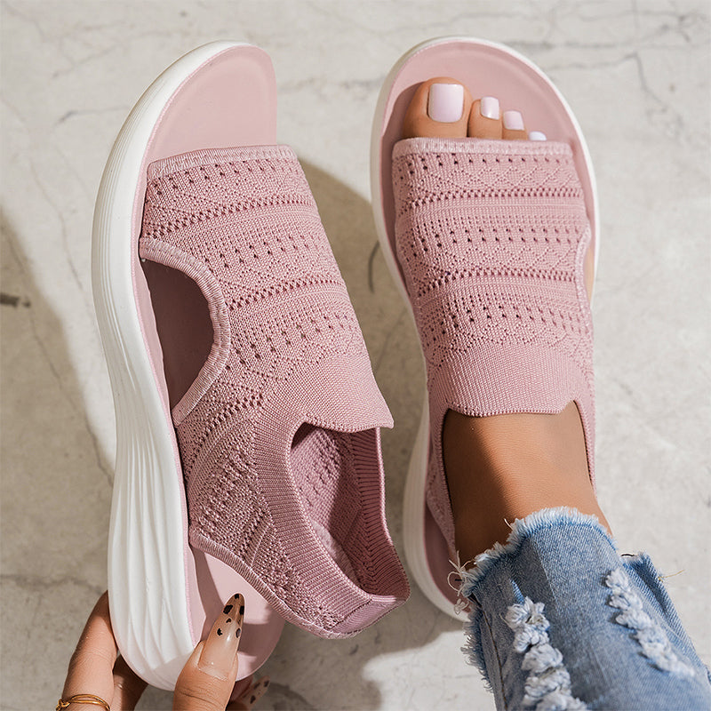 Pink breathable knit platform sandals with white soles on feet, one hand adjusting, on textured floor with distressed jeans.
