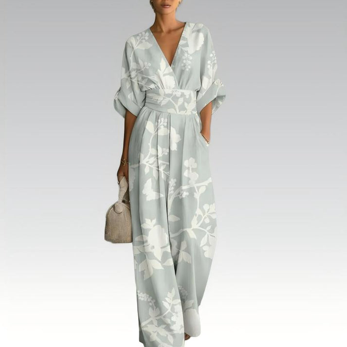 Light-colored jumpsuit with subtle leaf prints, deep V-neck, flowing design, accessorized with a handbag, on a neutral gradient backdrop.