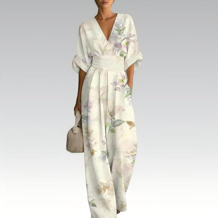 White jumpsuit with watercolor floral design, deep V-neck, cinched waist, holding a textured handbag, against a neutral gradient background.