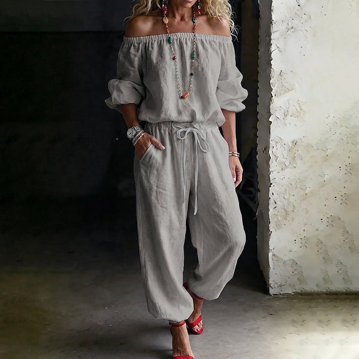 Woman in a gray off-shoulder relaxed jumpsuit with drawstring, indoors with textured walls.