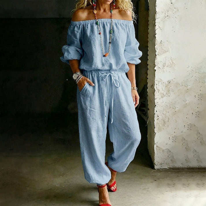 Woman in a blue off-shoulder relaxed jumpsuit with a drawstring waist, against an industrial indoor backdrop.