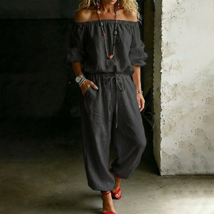Model in a black off-shoulder relaxed jumpsuit with drawstring, in a minimalistic indoor area.