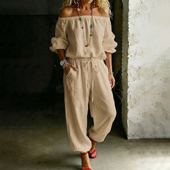Person in a beige off-shoulder relaxed jumpsuit with drawstring, in a textured wall indoor area.