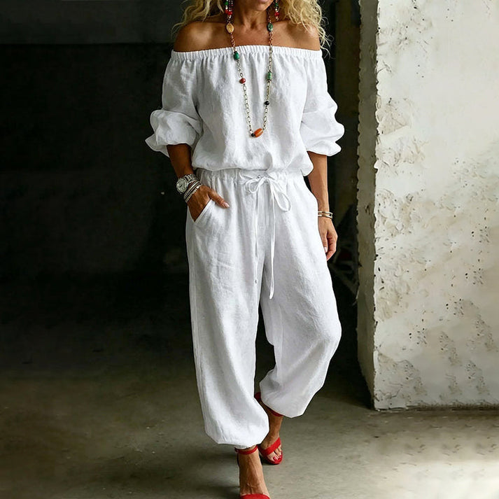 Woman wearing a white off-shoulder relaxed jumpsuit with drawstring, standing in an industrial indoor setting.