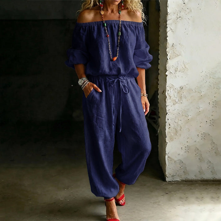 Model wearing a dark blue off-shoulder relaxed jumpsuit, featuring drawstring, in an indoor setting.