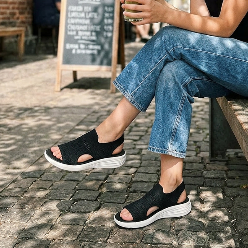Black knit platform sandals with white soles worn outdoors, person seated on a bench, cobblestone street setting.