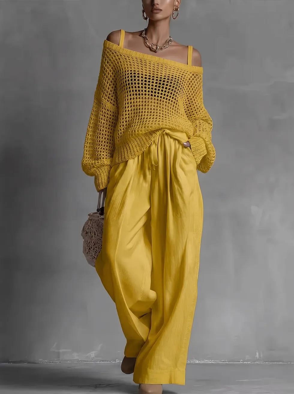 Woman in off-shoulder yellow top and wide yellow pants, holding woven bag in minimal gray studio.