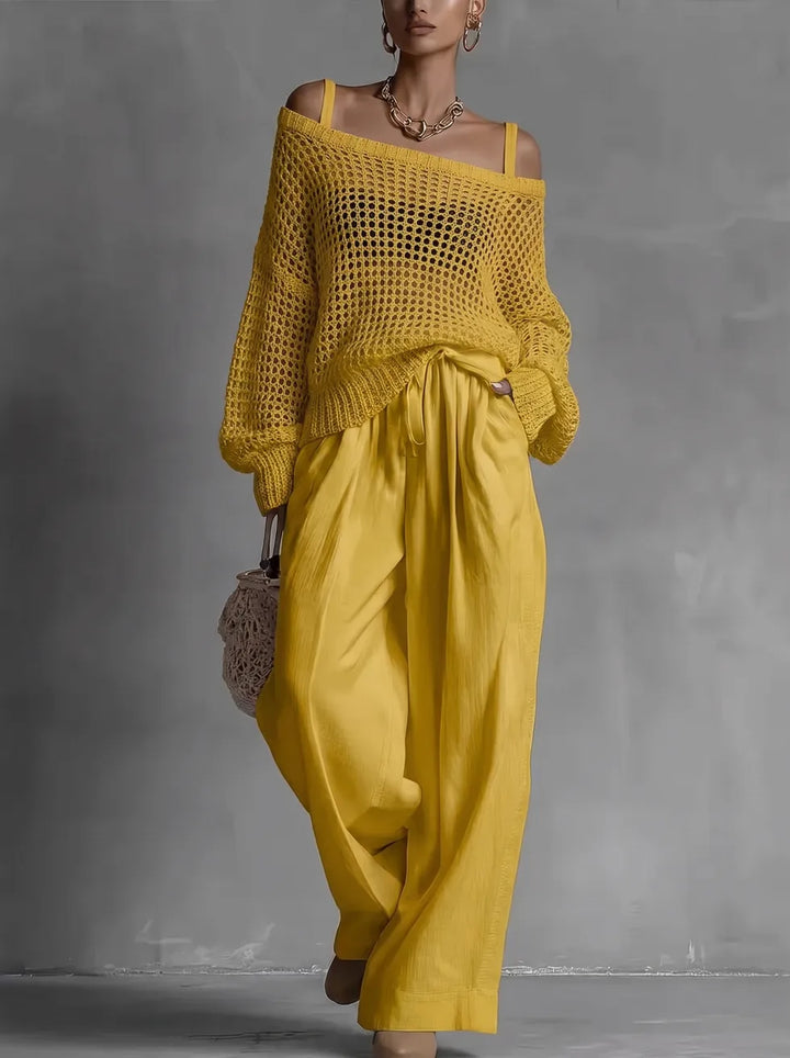 Woman in a yellow, off-shoulder knit top and matching flowing trousers, holding a netted bag, in a gray studio setting.