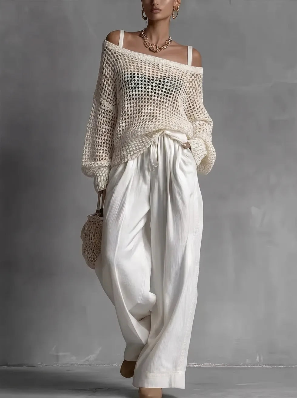 Woman in off-shoulder cream top and wide white pants, holding woven bag in minimal gray studio.