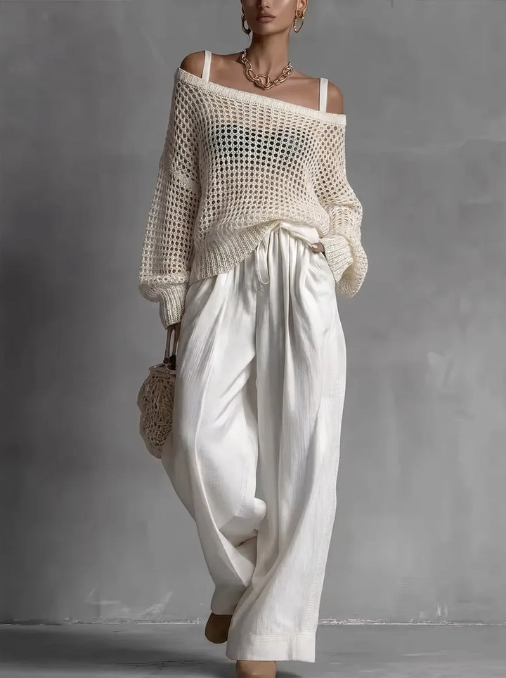 Woman in an ivory, off-shoulder knit top and flowing white trousers, holding a netted bag, in a gray studio setting.