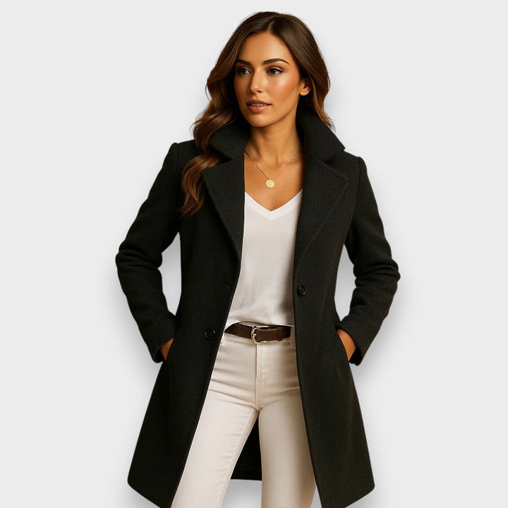 Model wearing a black classic coat over a white top and beige pants, studio background.