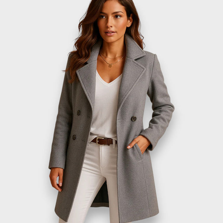 Model in a gray classic coat paired with a white top and beige pants, studio background.