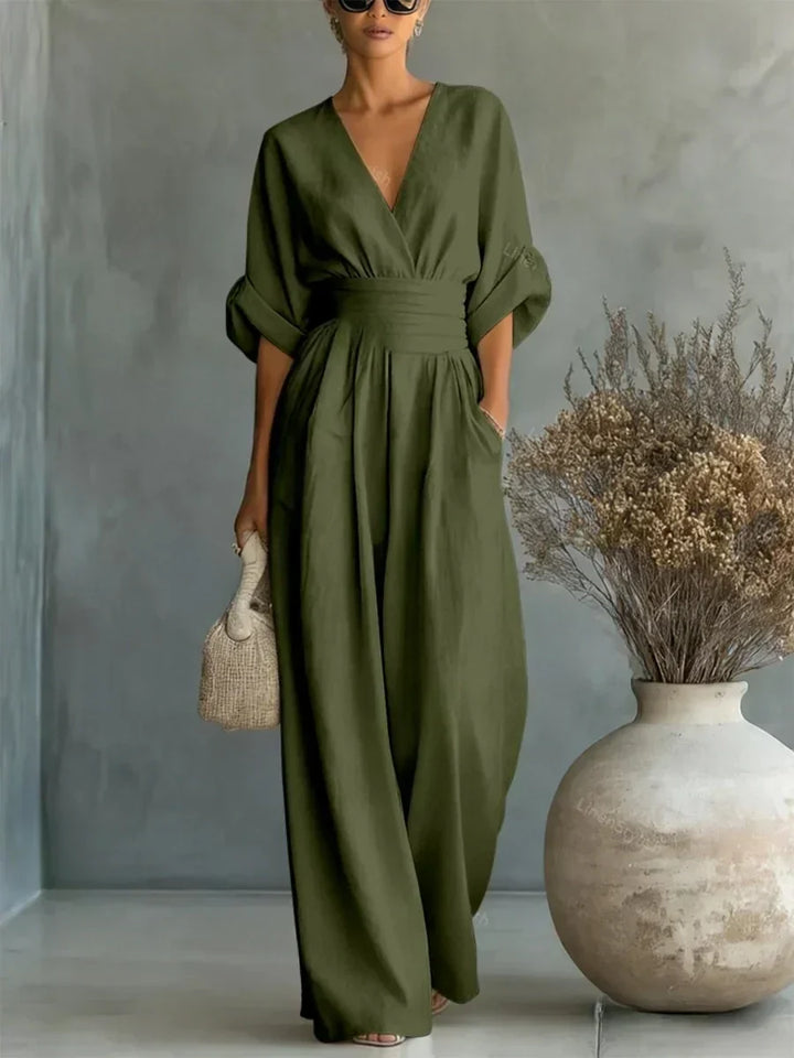 Olive green V-neck high-waist jumpsuit with rolled-up sleeves, presented in a studio with a decorative vase and subtle background.