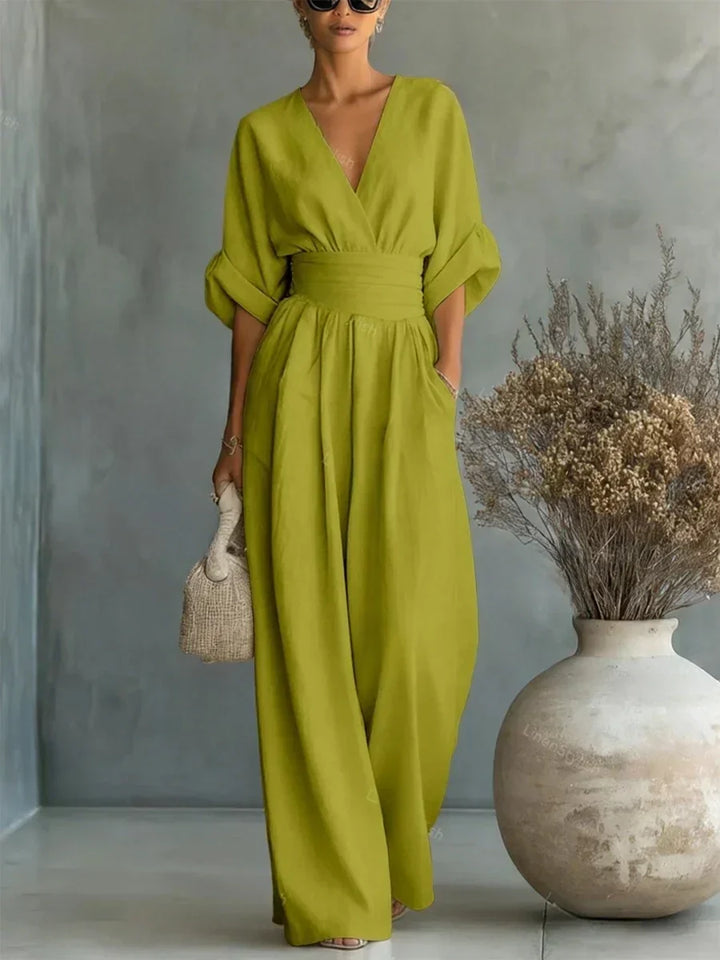 Lime green V-neck high-waist jumpsuit with rolled-up sleeves, modeled in a studio with a decorative vase and dried foliage.