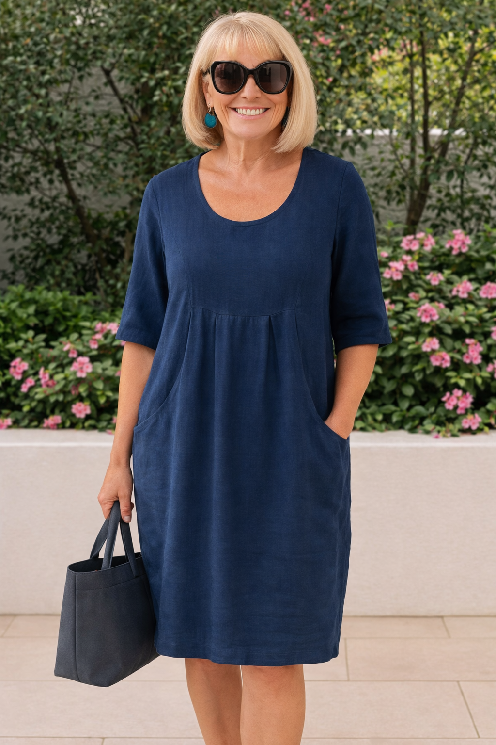 Woman in dark blue casual dress with pockets, standing outdoors with greenery and flowers, holding a dark bag.