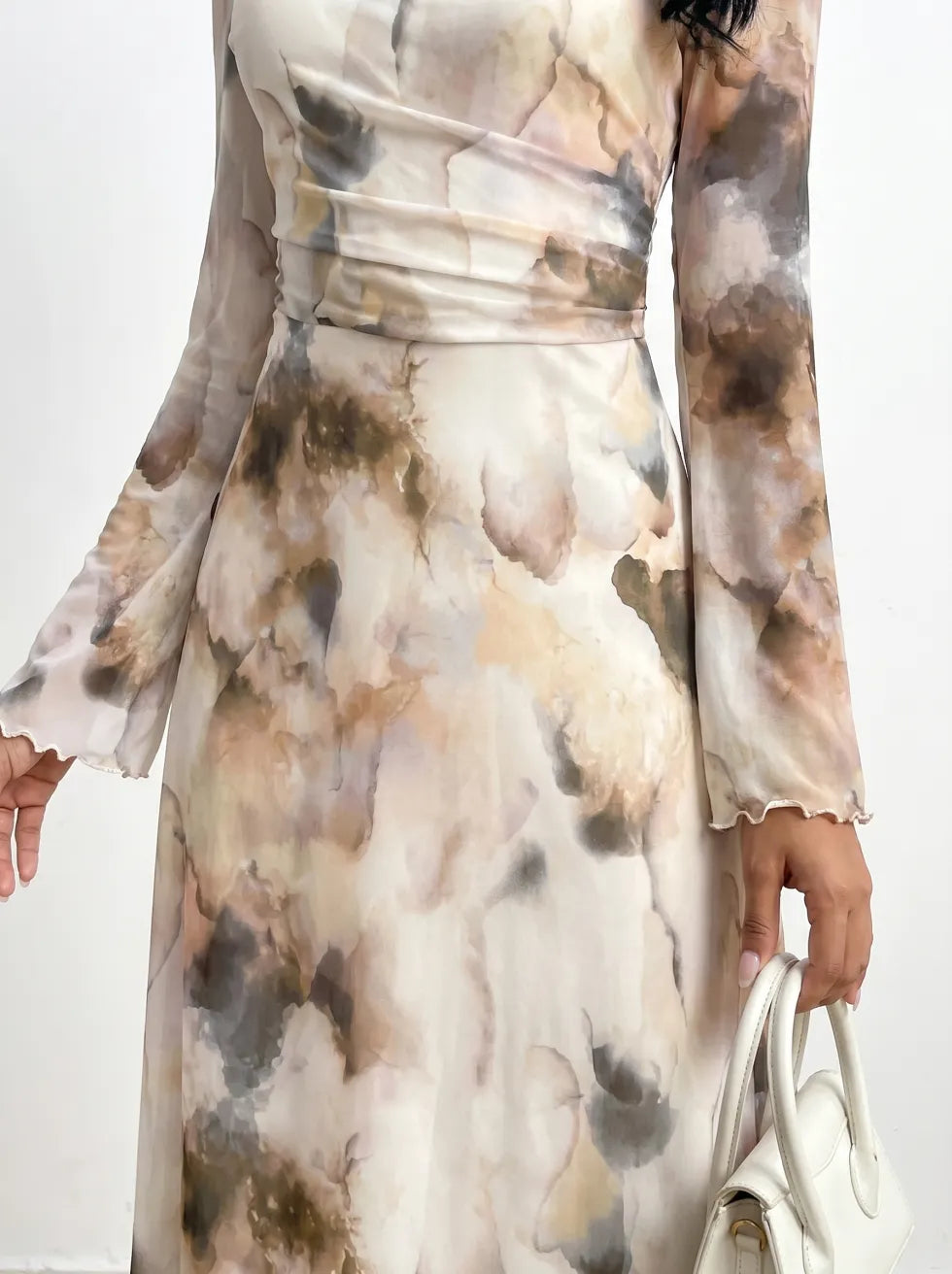 Detailed view of dress with an abstract watercolor design, cinched waist, flared sleeves, hand holding a white handbag, indoor background.