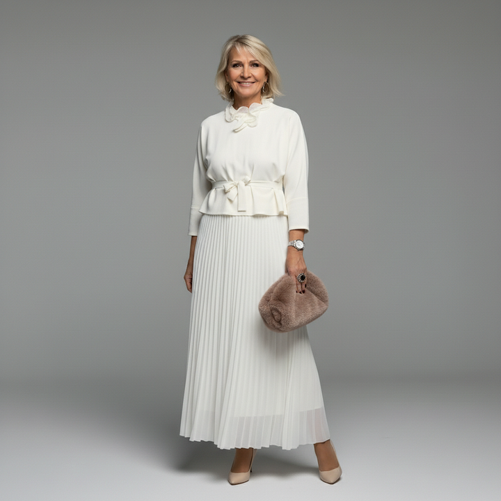 Woman in elegant white two-piece set with pleated skirt and ruffled top, holding beige clutch, studio background.