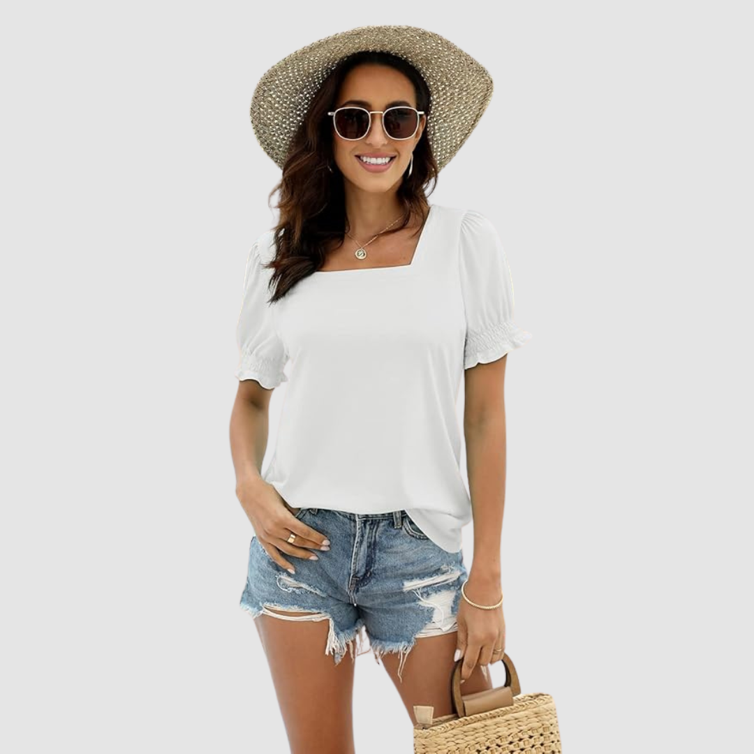 Woman in white square neck top, distressed denim shorts, straw hat and bag, studio background.
