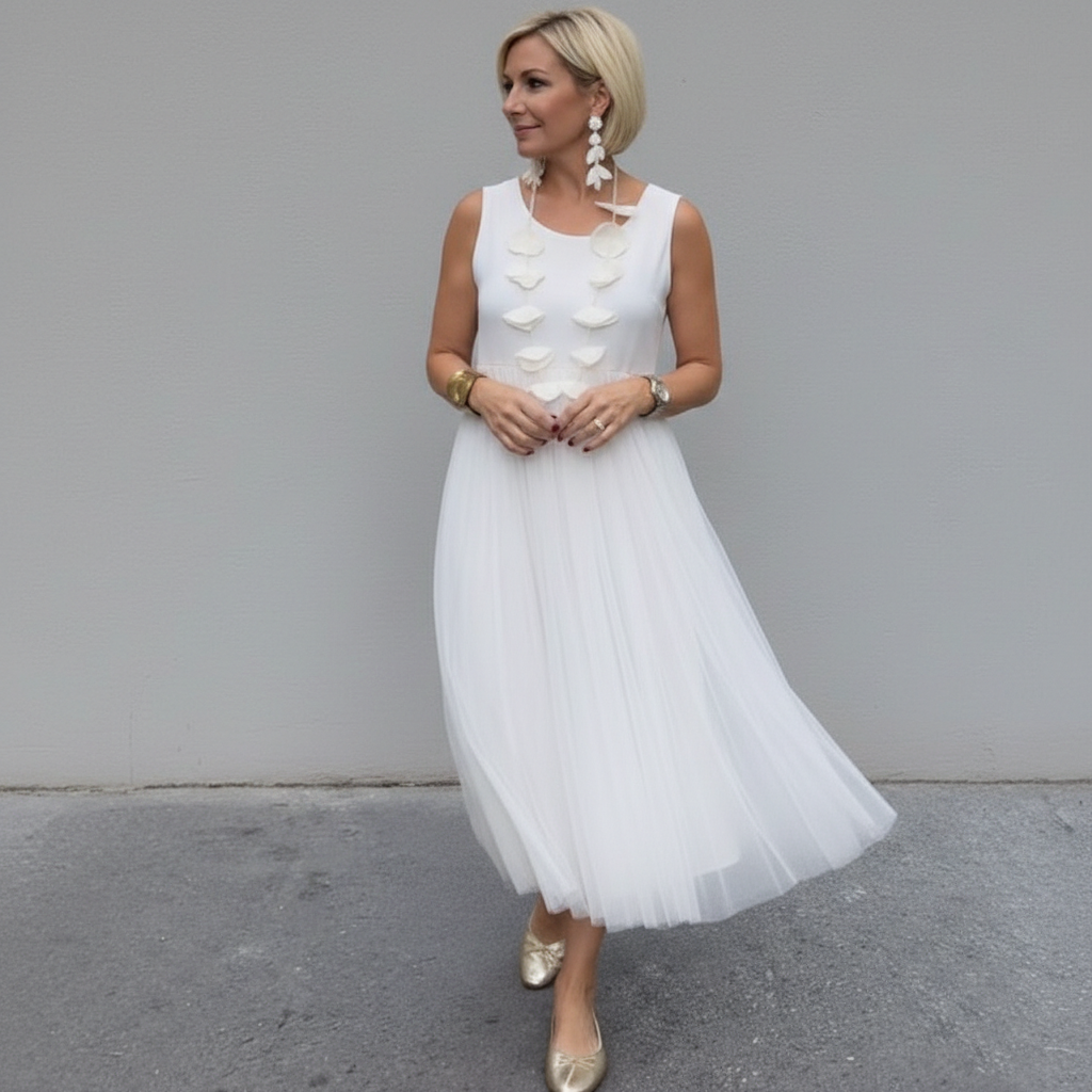 Woman in white pleated dress with statement necklace, standing outdoors by a gray wall.
