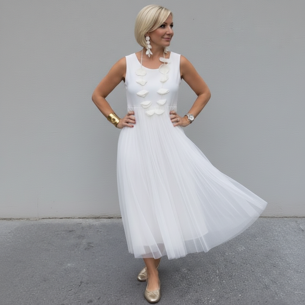 Elegant woman in white pleated dress with a necklace, posing outdoors with a gray background.