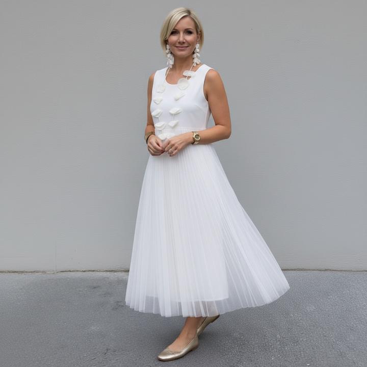 Woman in white pleated dress with statement necklace, standing against a gray wall outdoors.